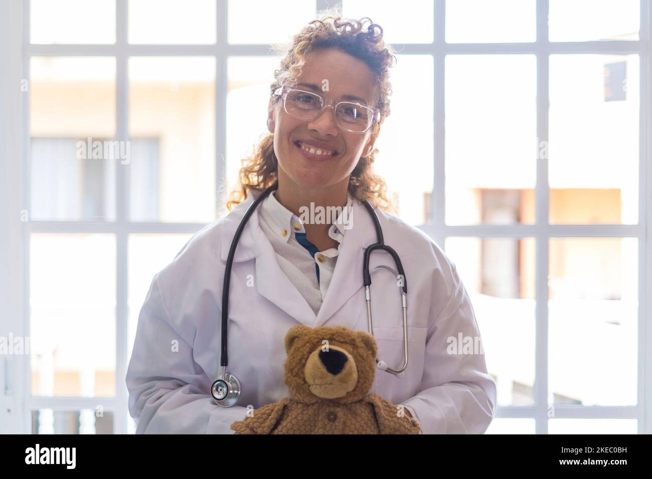 Portriat de jeune femme médecin en uniforme et stéthoscope tenant l'ours en peluche contre la fenêtre à la clinique. Professionnel de la santé ou professionnel de la santé confiant dans les lunettes à l'hôpital Banque D'Images