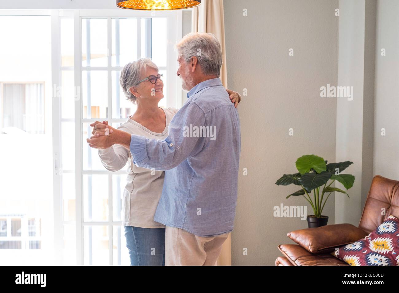 Couple sénior romantique tenant les mains en appréciant danser ensemble dans le salon de la maison, couple heureux âgé célébrant en dansant dans le salon. Banque D'Images