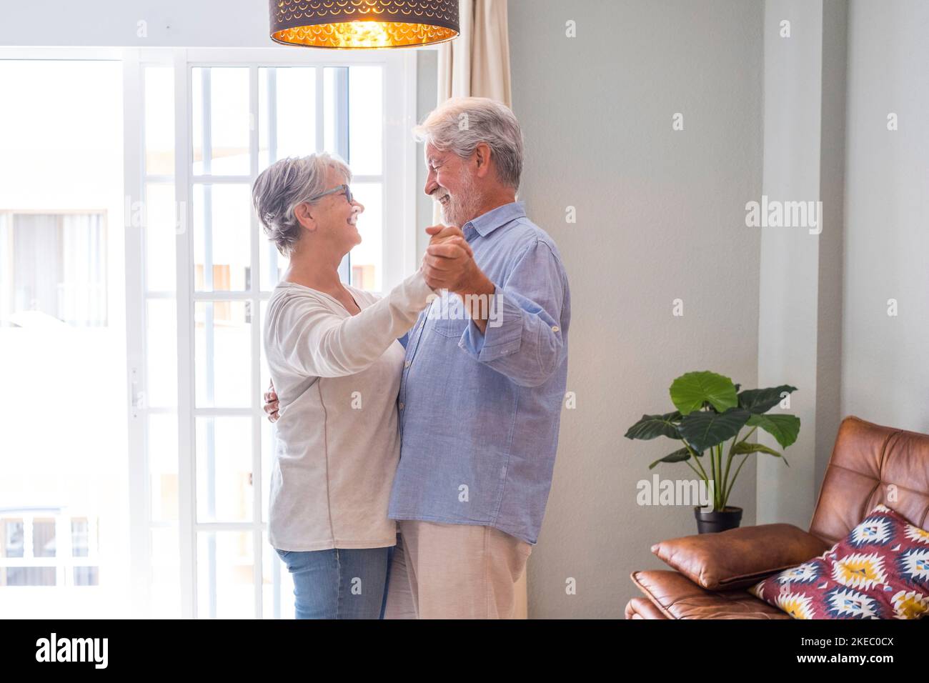 Couple sénior romantique tenant les mains en appréciant danser ensemble dans le salon de la maison, couple heureux âgé célébrant en dansant dans le salon. Banque D'Images