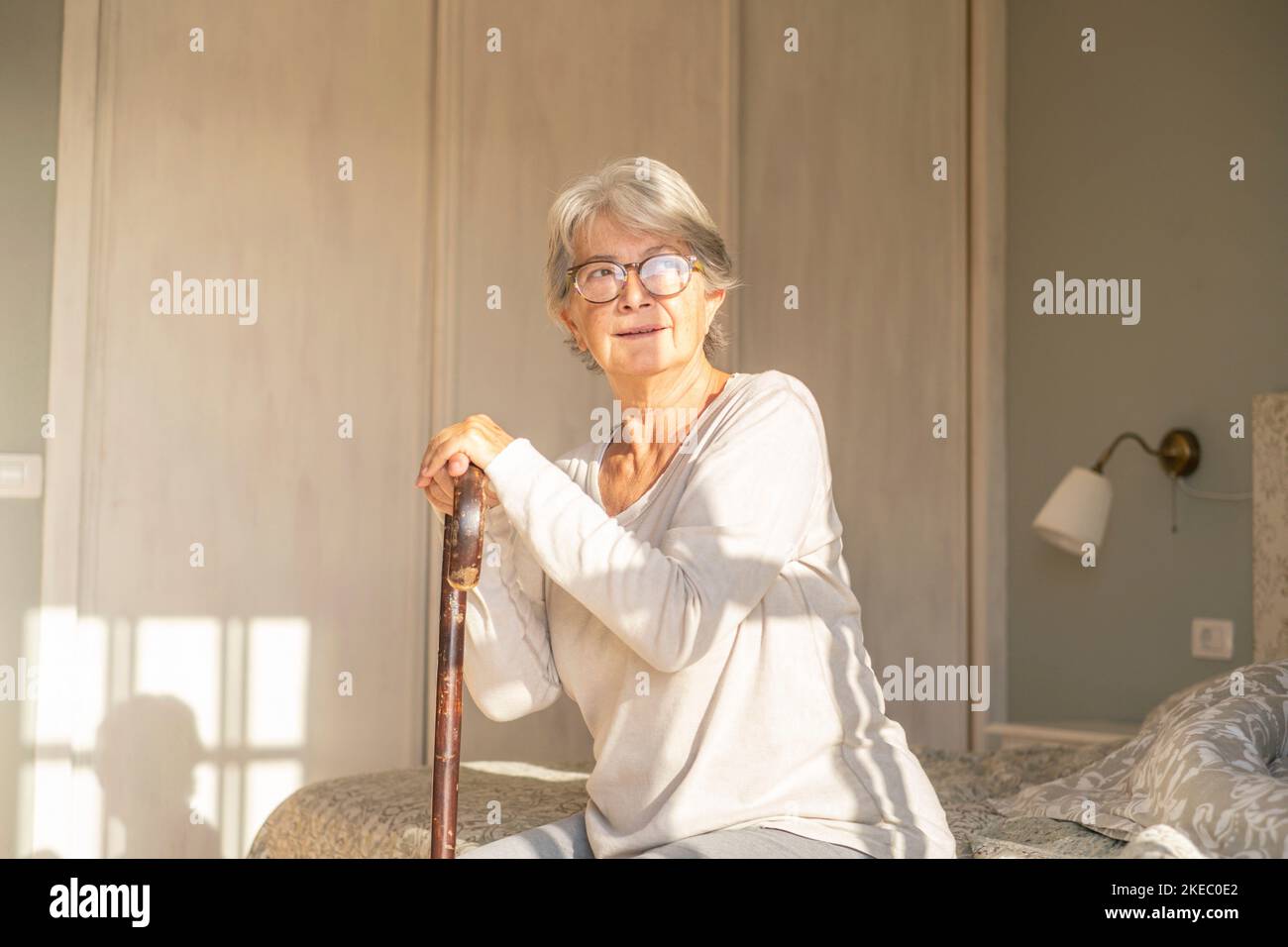 Femme séchée sur un bâton de marche tout en étant assise sur le lit. Femme solitaire âgée assise dans la chambre à la maison. Une vieille femme qui profite de la lumière du soleil matinale à la maison Banque D'Images