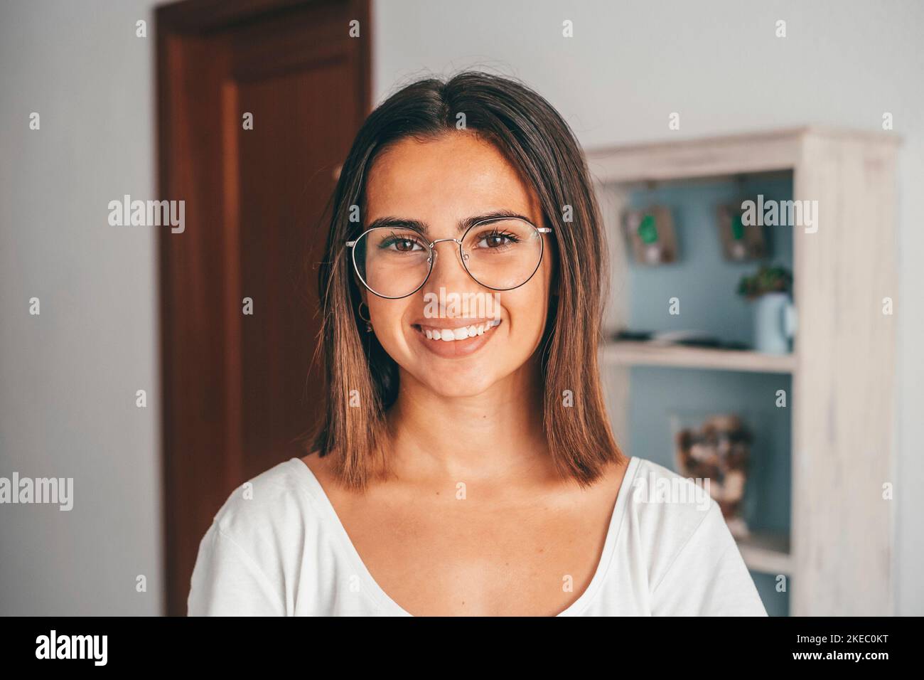 Bonne jeune femme en lunettes regardant l'appareil photo de la maison de sa maison. Belle dame satisfaite avec des cheveux rabots souriant en faisant face à l'appareil photo dans un appartement moderne. Jeunes femmes souriant à la maison Banque D'Images