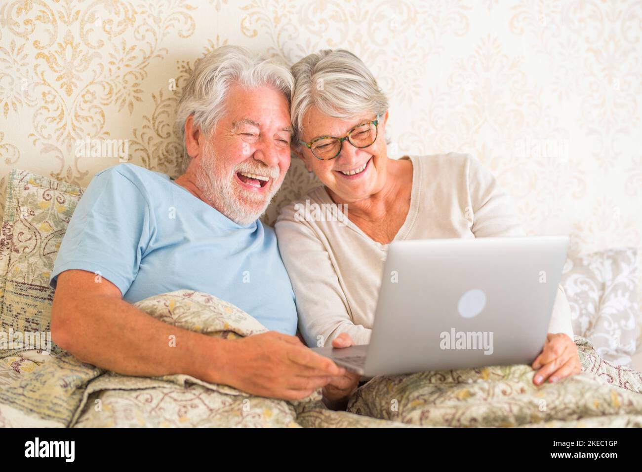 Un vieux couple caucasien riant et utilisant un ordinateur portable le matin au lit dans la chambre à la maison. Couple de personnes âgées surfant et utilisant les médias sociaux sur ordinateur portable à la maison. Banque D'Images
