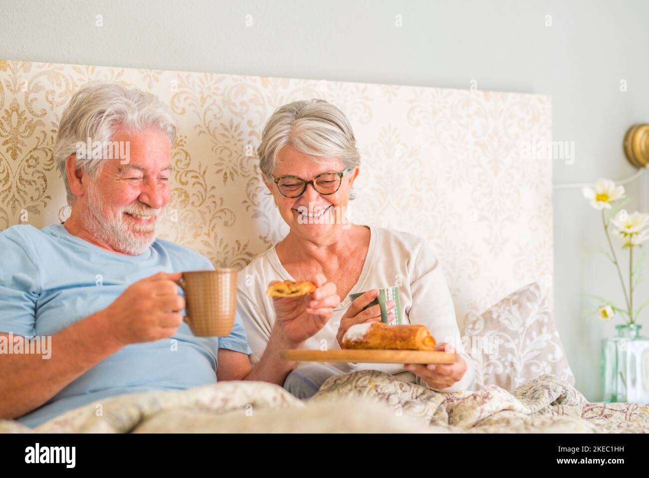 Un vieux couple caucasien prend le petit déjeuner le matin au lit dans la chambre à la maison. Couple de personnes âgées prenant un croissant et buvant du café de la tasse pour le petit déjeuner à la maison. Banque D'Images