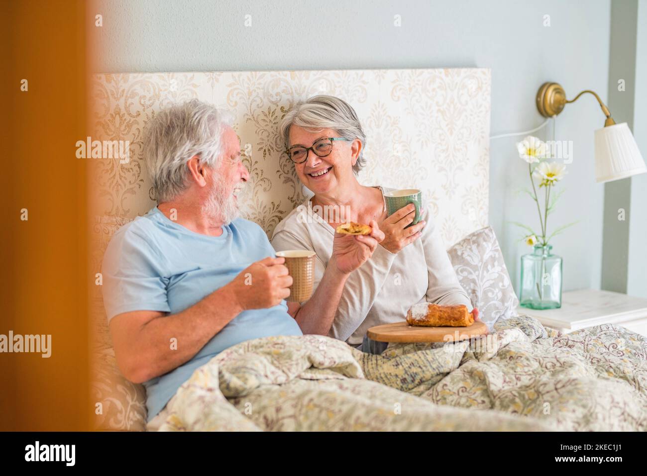 Un vieux couple caucasien riant et prenant le petit déjeuner le matin au lit dans la chambre à la maison. Couple de personnes âgées prenant un croissant et buvant du café de la tasse pour le petit déjeuner à la maison Banque D'Images