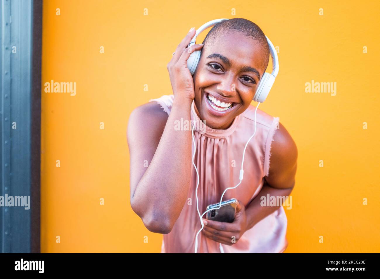 une jeune et belle femme africaine ou américaine souriante et regarder l'appareil photo tenant un casque sur sa tête écouter de la musique vibing et danser seul avec fond jaune - des gens heureux qui entendent de la musique Banque D'Images