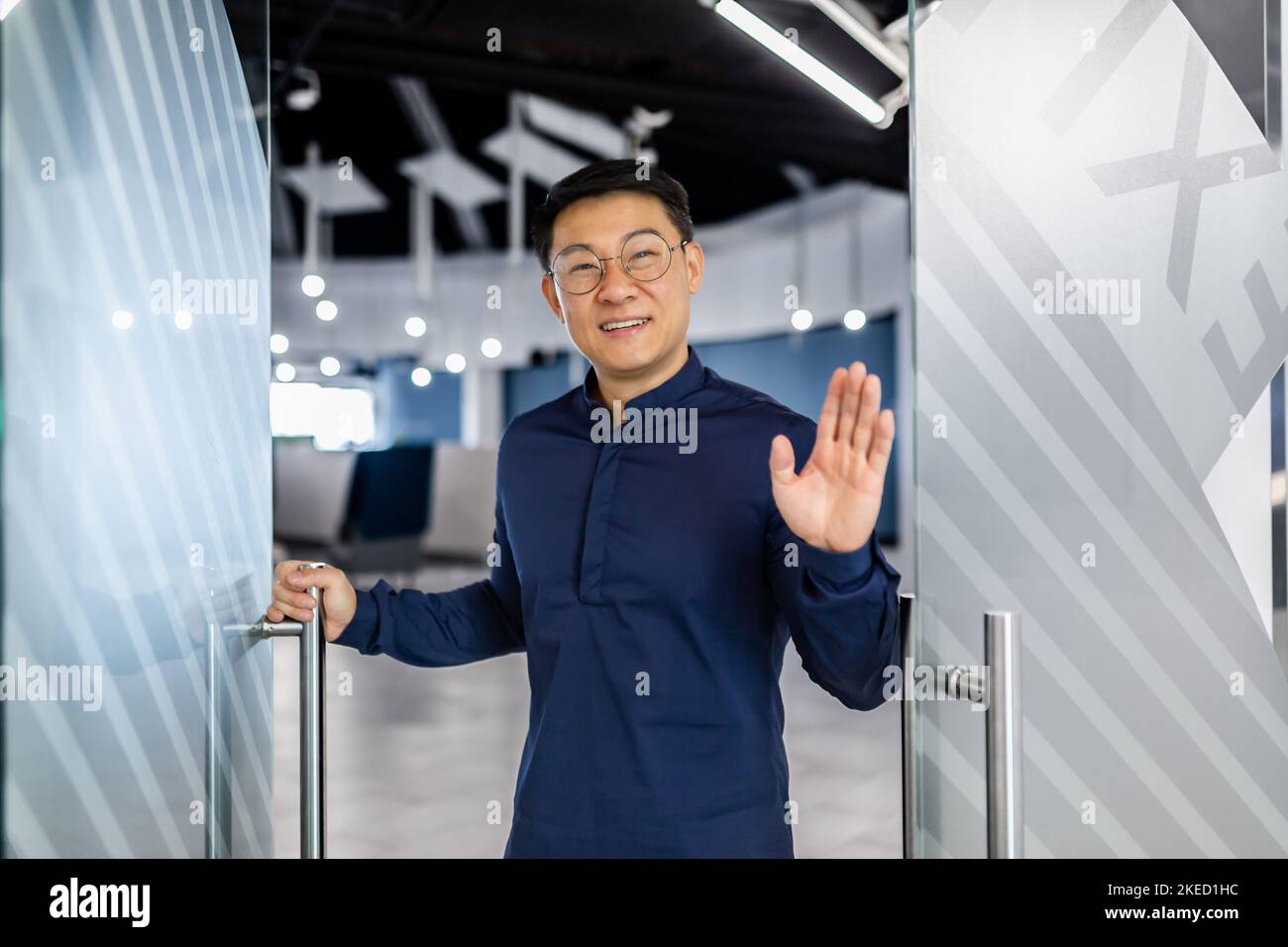 Un jeune beau architecte asiatique, concepteur, ingénieur se tient à la porte d'un bureau moderne, accueille, sourit à la caméra. Banque D'Images