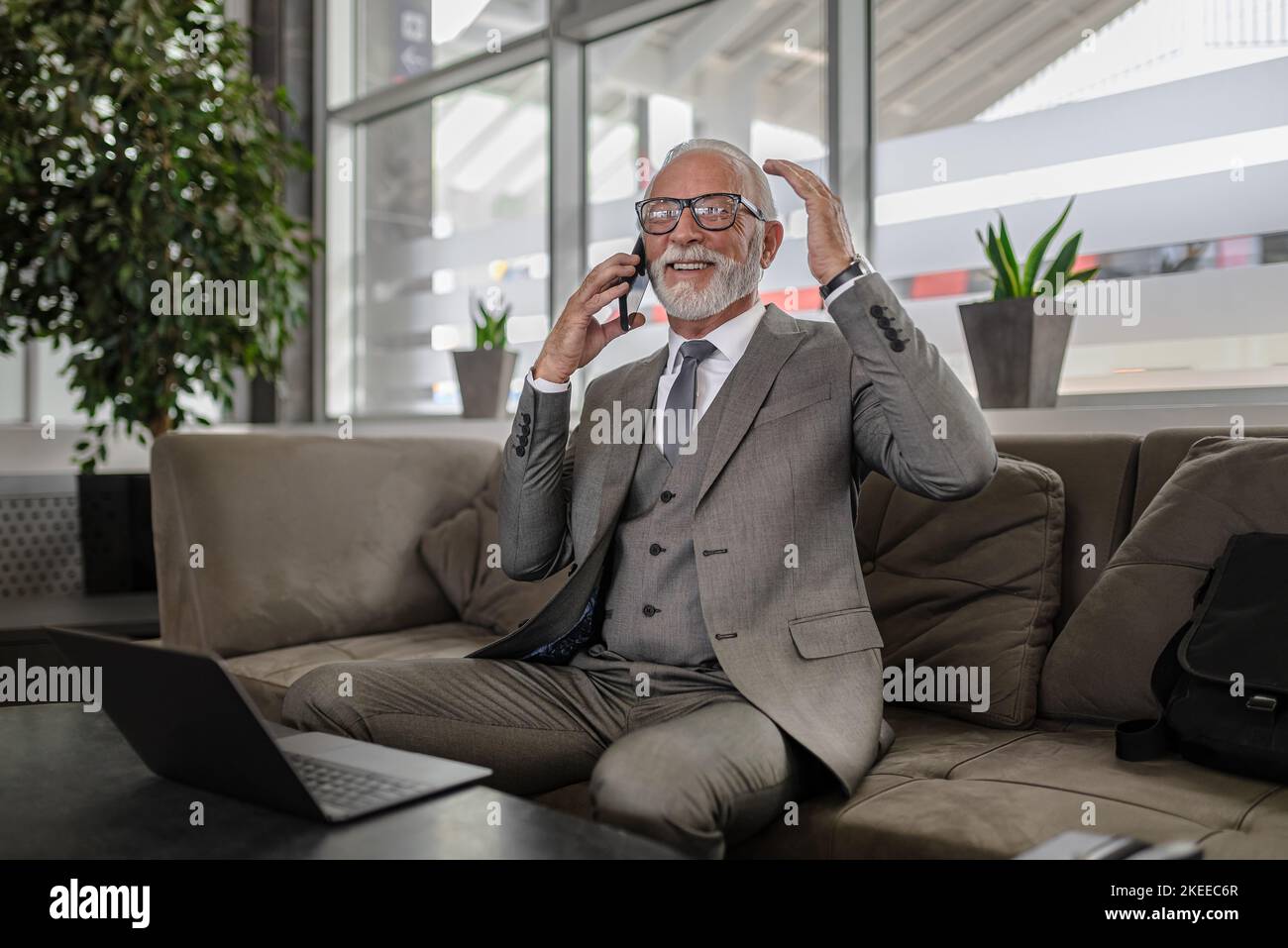 Souriant entrepreneur âgé faisant des gestes tout en discutant sur téléphone portable. Homme d'affaires senior utilise un ordinateur portable à table. Il porte un costume d'affaires Banque D'Images