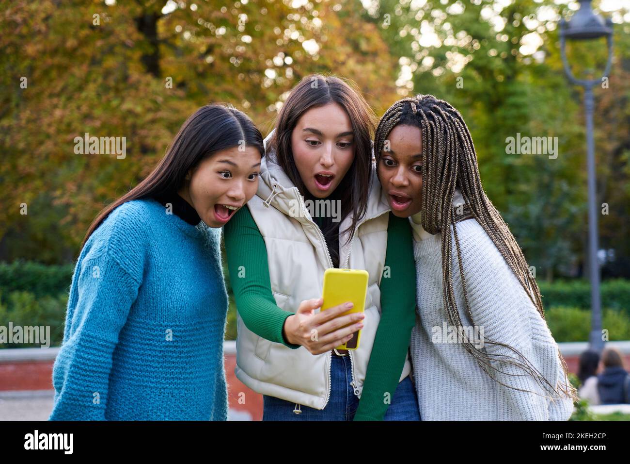 Trois femmes multiethniques émerveillement devant le téléphone portable, debout à l'extérieur du parc public de la ville. Banque D'Images