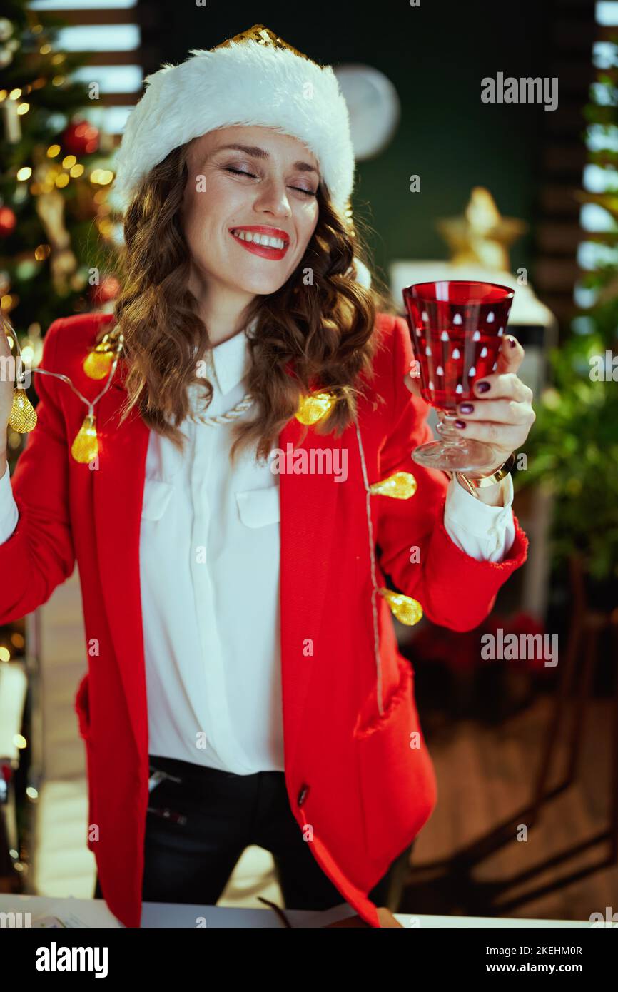 Noël. Souriante élégante femme d'affaires de 40 ans en chapeau de père noël et veste rouge avec danse de verre de vin dans un bureau moderne vert avec Noël t Banque D'Images