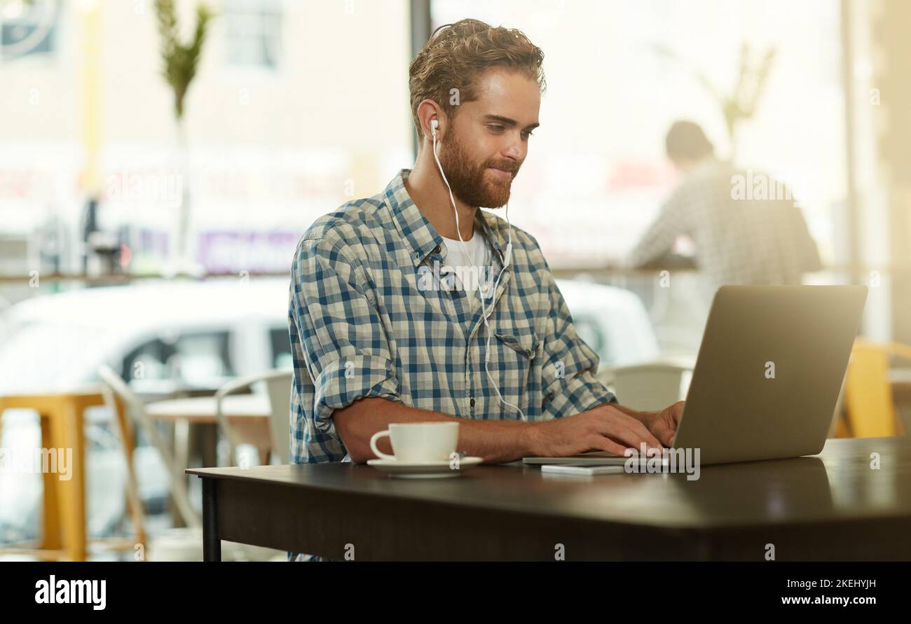 Il sait comment faire les choses. Un jeune homme avec des écouteurs utilisant un ordinateur portable dans un café. Banque D'Images
