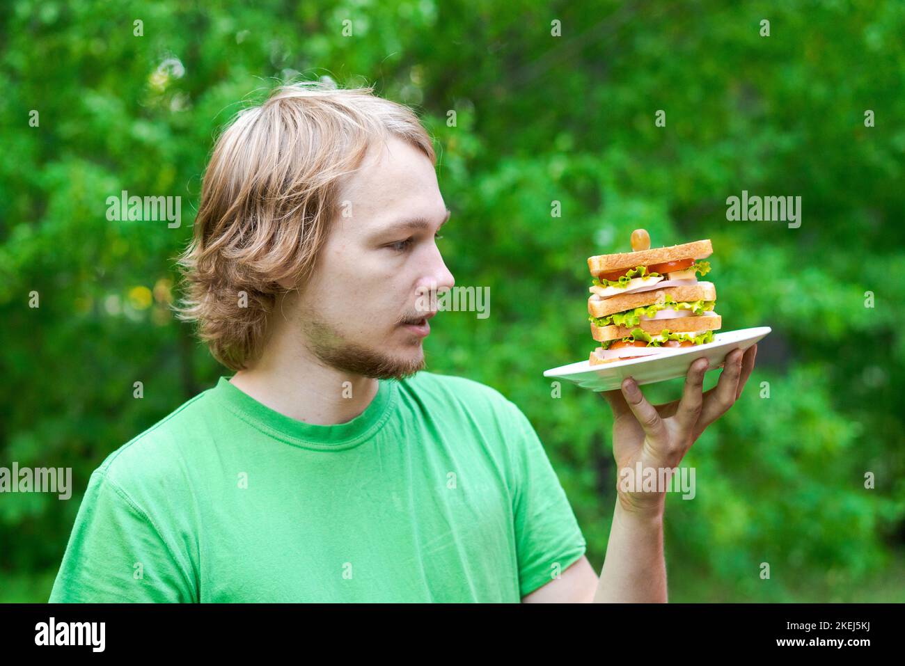 Jeune homme tenant un gros sandwich à la saucisse sur l'assiette. L'étudiant mange de la nourriture rapide dans un paquet sur fond de feuillage vert. Aliments indésirables rapides. Un homme très affamé. Concept de régime. Banque D'Images