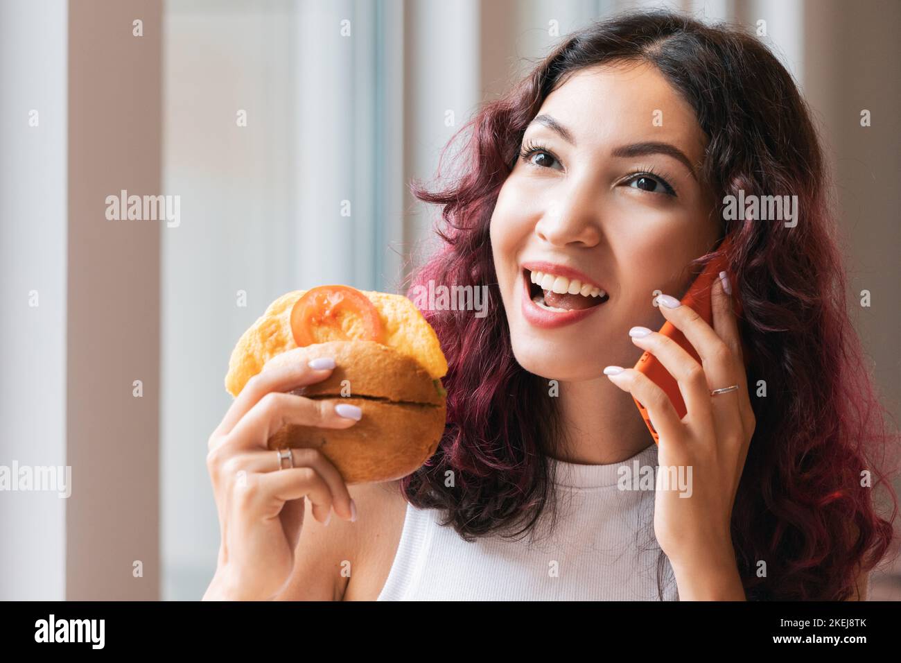Une femme mange un hamburger dans un fast-food café et parle à un ami au téléphone Banque D'Images