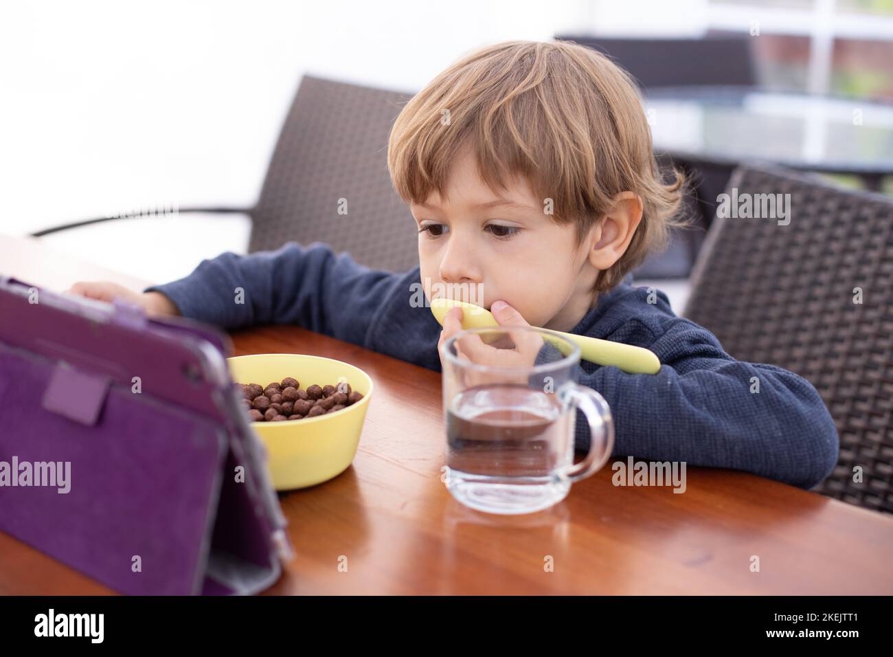 Un petit garçon s'assoit sur une chaise à la table et regarde le dessin animé sur une tablette, l'ordinateur, la cuillère à lécher. Les enfants de la maternelle mangent des boules de chocolat avec du lait et des boissons Banque D'Images