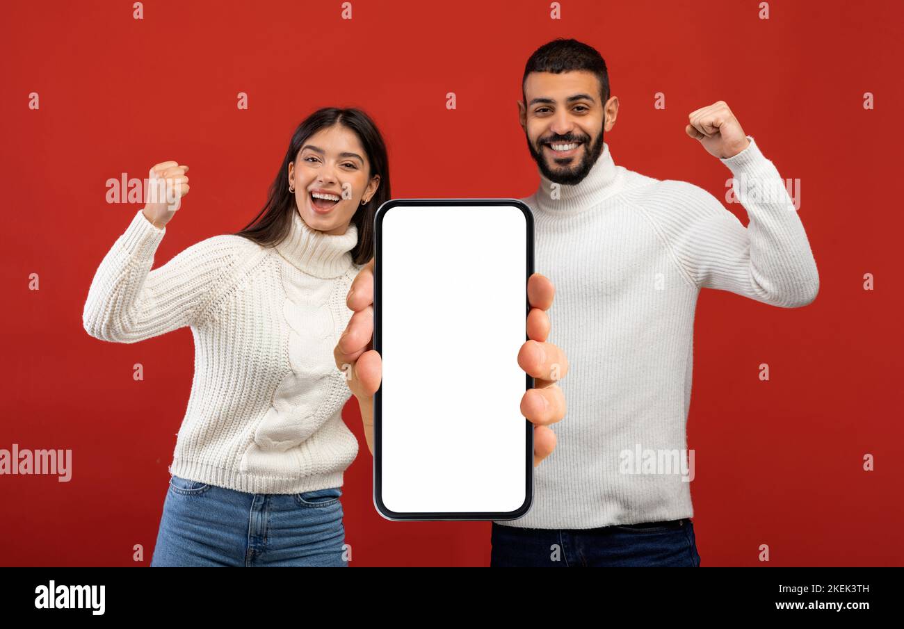 Joyeux couple arabe millénaire heureux fêtez la victoire avec des mains gestuelles, montrez un grand smartphone avec un écran vierge Banque D'Images