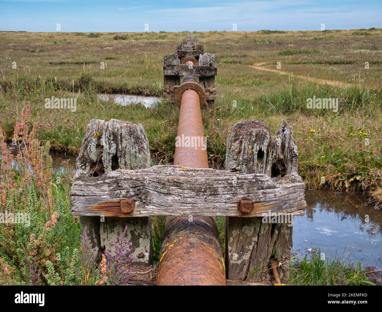 Un tuyau de drainage rouillé sur des supports en bois altérés aux marais de sel de raidisseur sur le chemin de la côte nord de Norfolk. Le tuyau traverse les marais. Banque D'Images