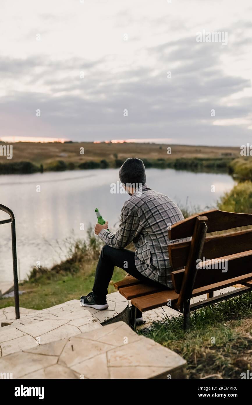 Un jeune homme s'assoit sur un banc sur les rives d'une rivière, d'un lac ou d'une baie et boit de la bière. Pique-nique d'automne dans le parc. Feuilles jaunes et repos. Banque D'Images