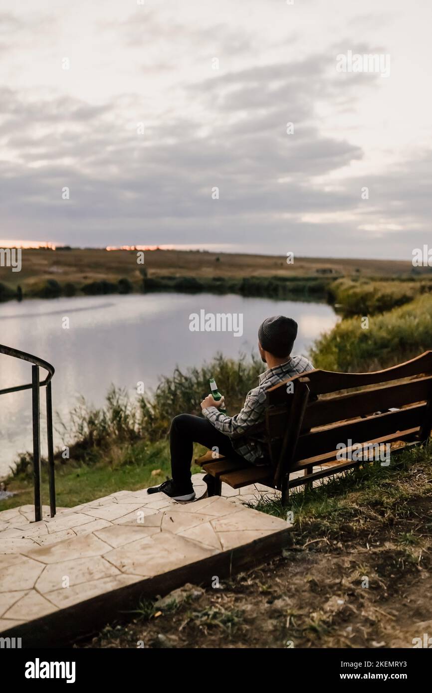 Un jeune homme s'assoit sur un banc sur les rives d'une rivière, d'un lac ou d'une baie et boit de la bière. Pique-nique d'automne dans le parc. Feuilles jaunes et repos. Banque D'Images