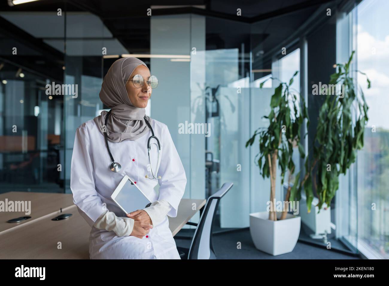 Une jeune femme musulmane heureuse et réfléchie, médecin arabe à l'hôpital, est assise à son bureau, tenant une tablette entre ses mains, regardant la fenêtre, souriant, aimant son travail. Banque D'Images