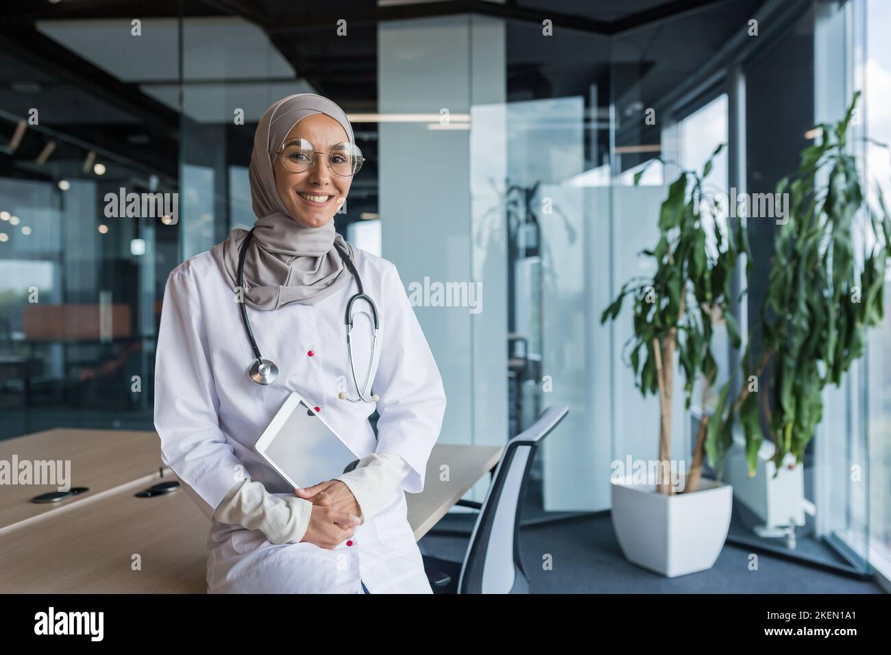 Portrait d'une jeune femme arabe musulmane heureuse et belle, étudiante assise sur une table dans un grand bureau dans un hôpital, tenant une tablette entre ses mains, regardant la caméra, souriant. Banque D'Images