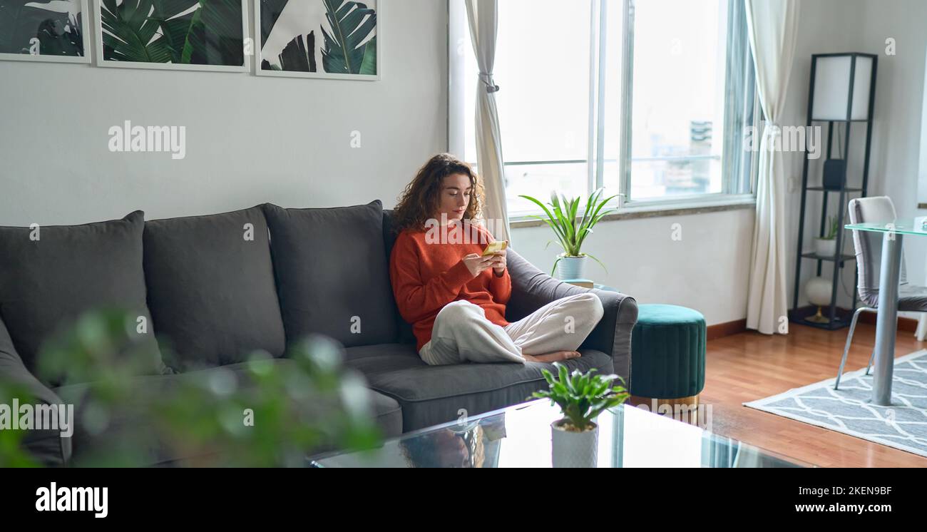 Jeune femme utilisant un téléphone portable assis sur un canapé dans la salle de séjour. Banque D'Images