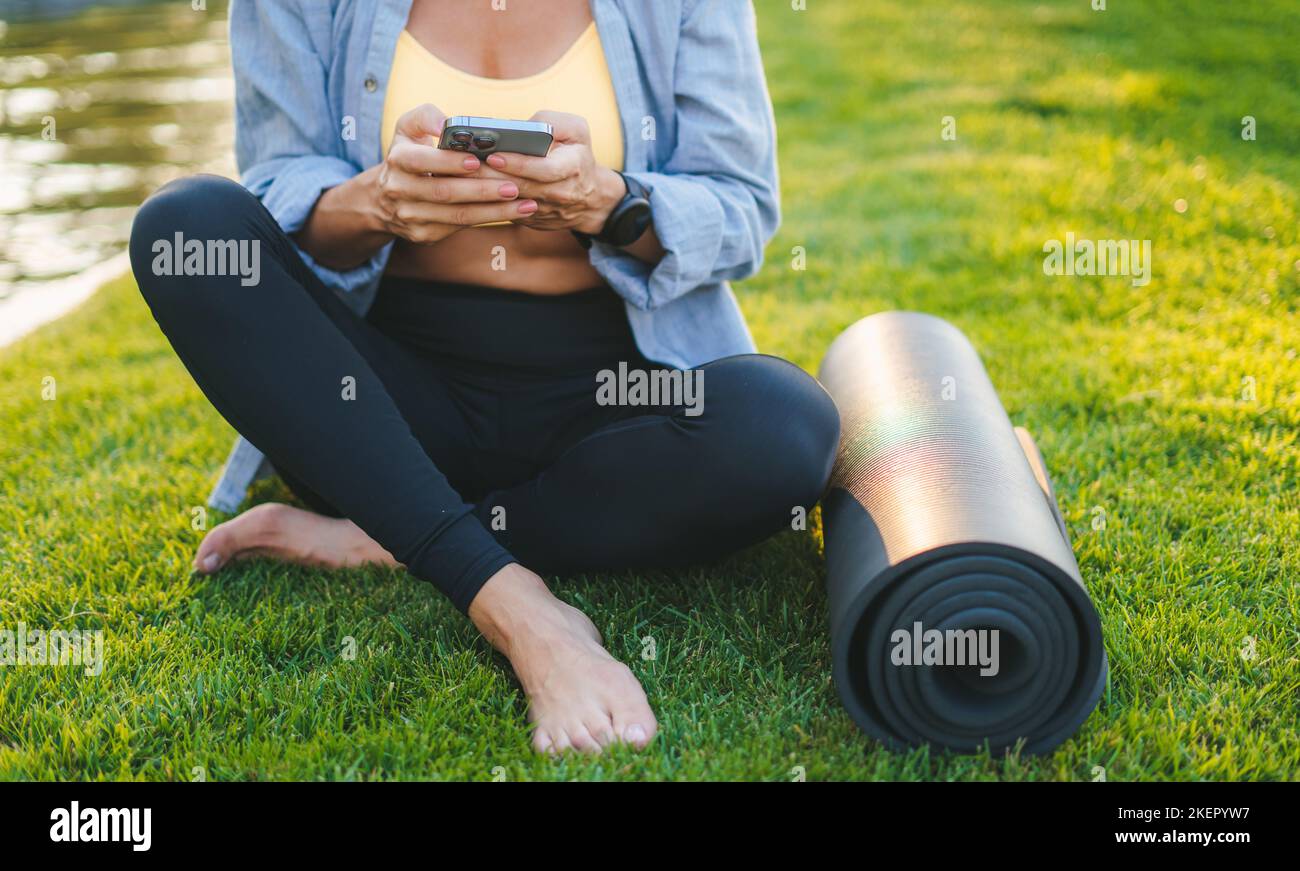 Femme sportive tenant le téléphone assis sur l'herbe verte près du tapis de yoga, se reposant après l'entraînement du matin. Entraînement. Femme de fitness. En bonne santé Banque D'Images