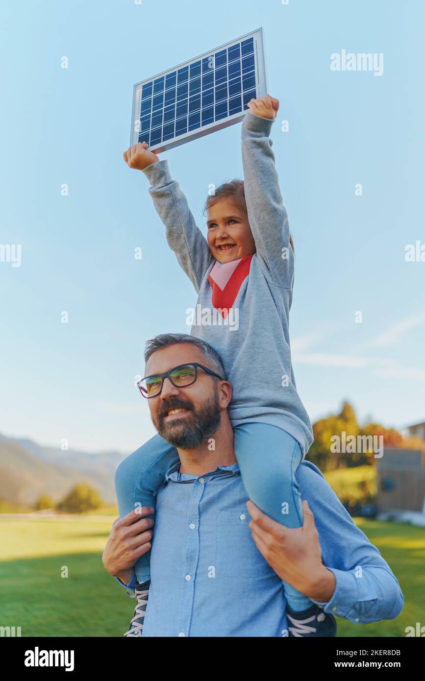 Père avec sa petite fille sur le porcgyback, prenant le soleil au panneau solaire, le chargeant à leur arrière-cour. Énergies alternatives, économies de ressources et Banque D'Images