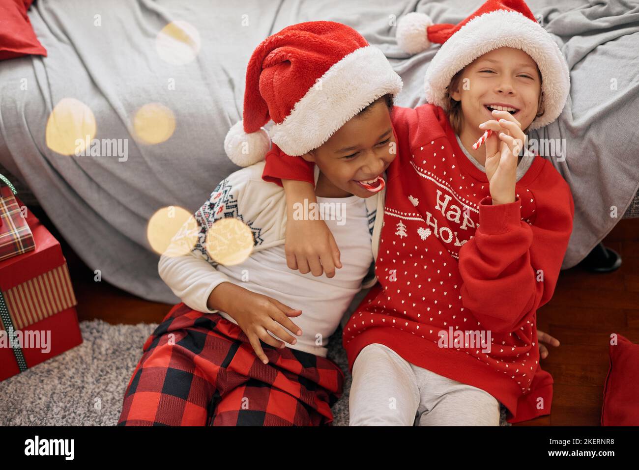 Joyeux garçons interraciaux portant des costumes de fête, des chapeaux de Père Noël, des cannes de bonbons de lécher, s'amuser Banque D'Images