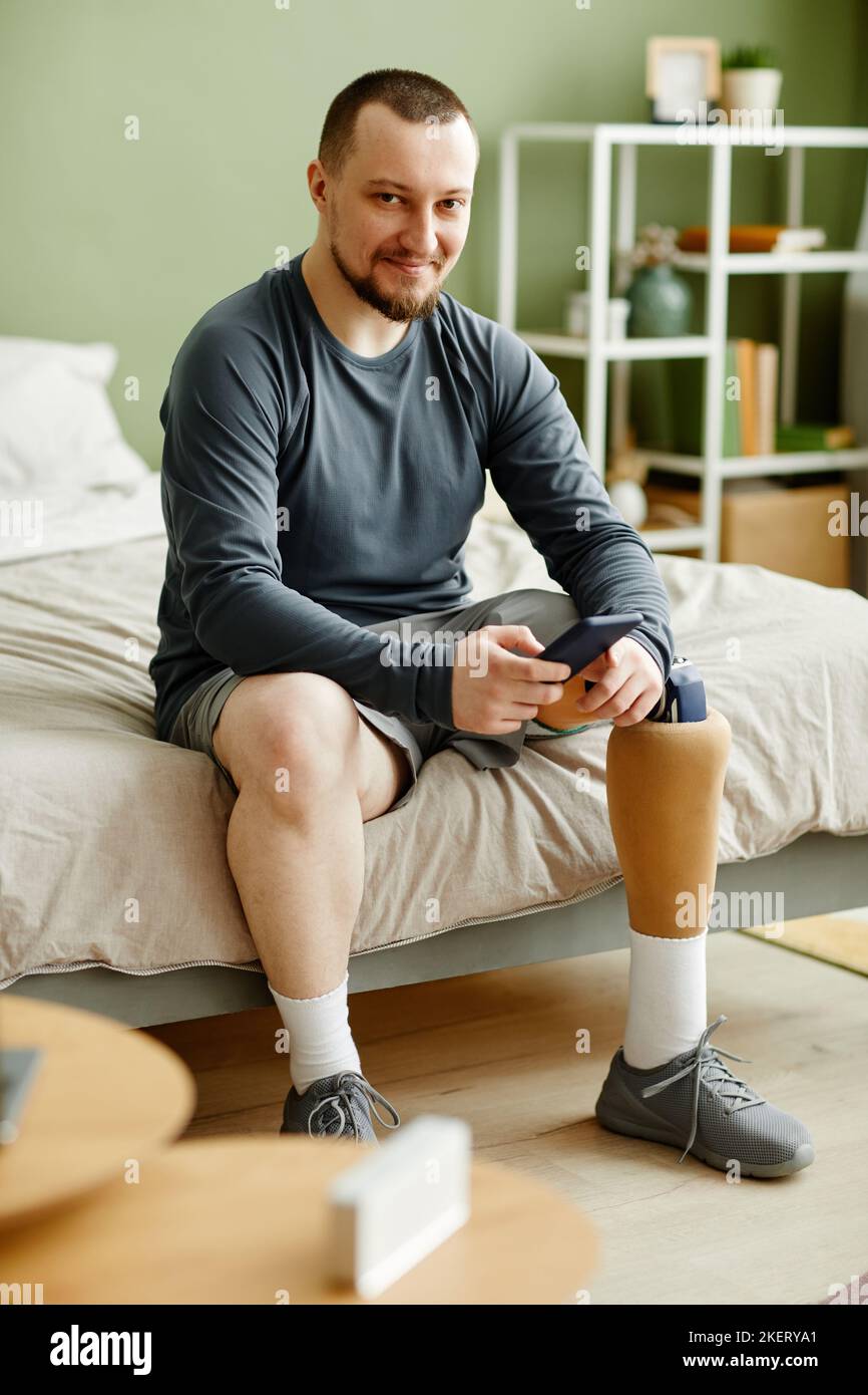 Portrait vertical complet d'un homme avec une jambe prothétique utilisant un smartphone et souriant à la caméra tout en étant assis sur le lit à la maison Banque D'Images