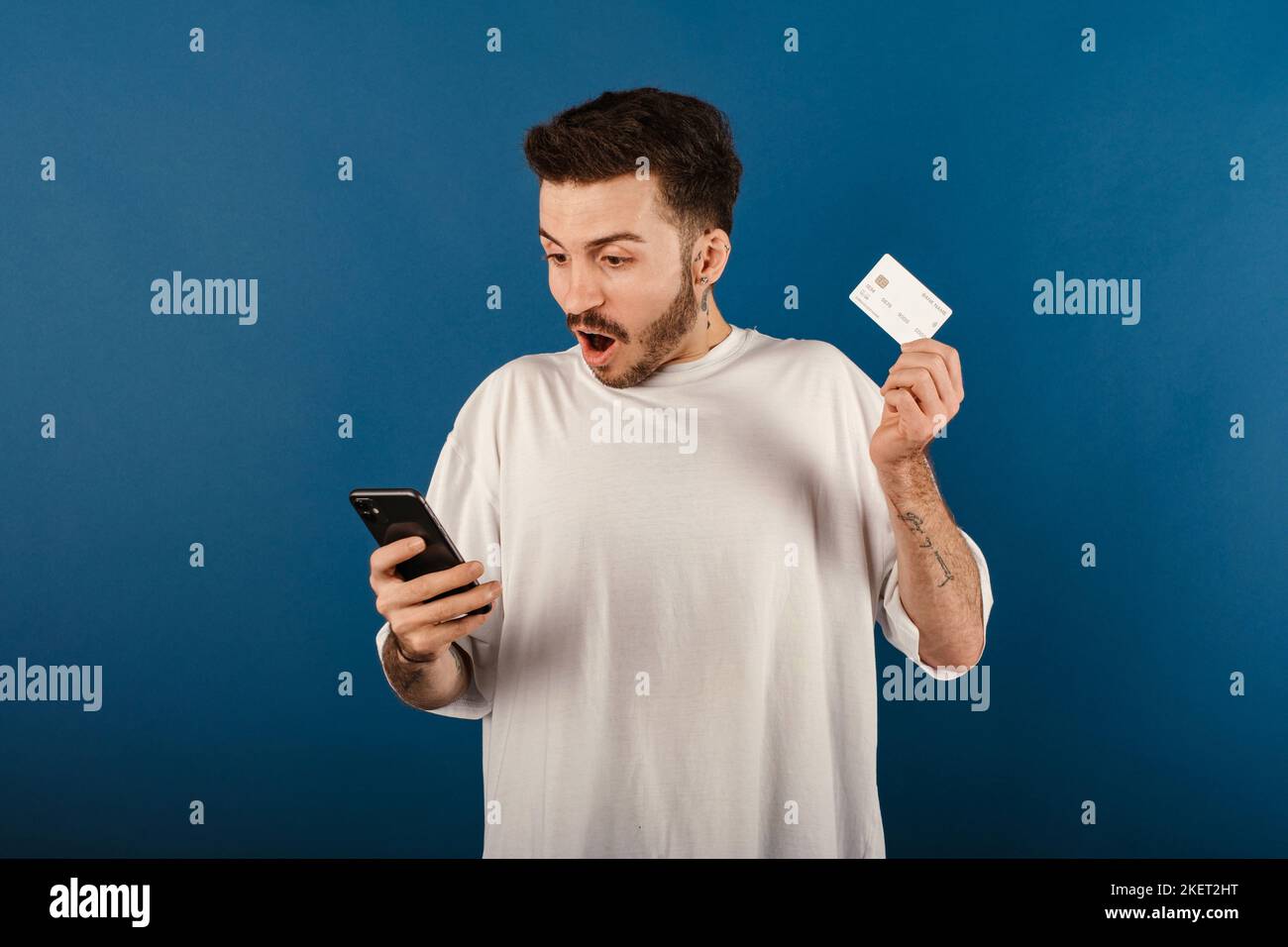Portrait d'un homme joyeux portant un t-shirt blanc posant isolé sur fond bleu tenant un smartphone et une carte de crédit choqué de surprise et d'émerveiller Banque D'Images