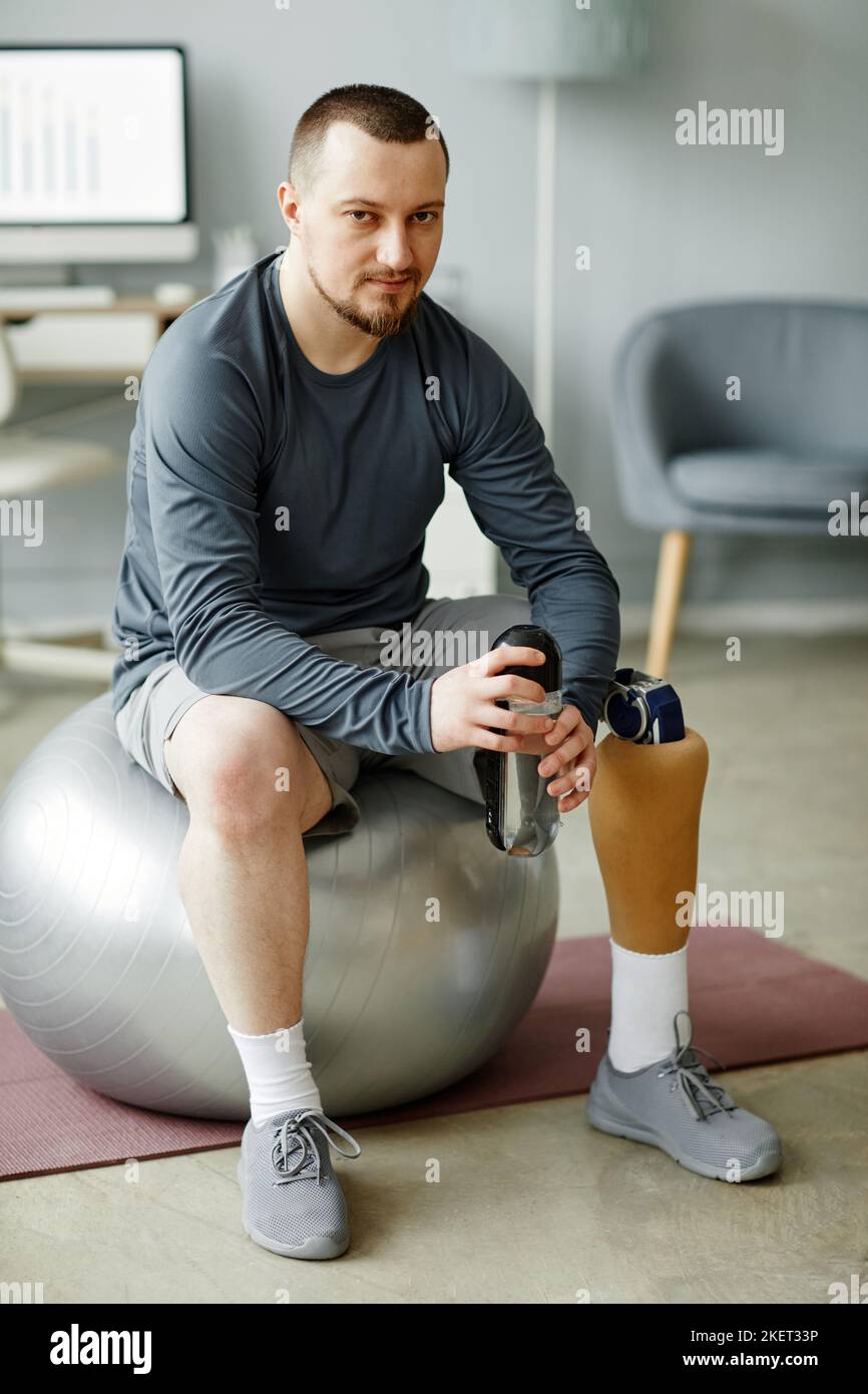 Portrait vertical complet d'un homme avec une jambe prothétique assis sur le ballon de fitness et tenant une bouteille d'eau après l'entraînement à la maison Banque D'Images