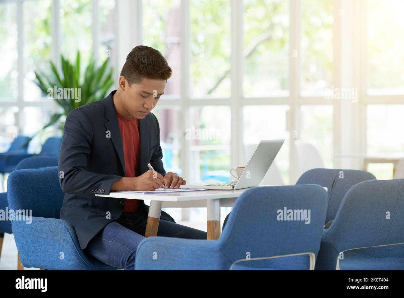 Jeune entrepreneur concentré assis dans un grand hall de bureau avec fenêtres panoramiques et prenant les notes nécessaires tout en préparant les négociations importantes, portrait-shot Banque D'Images