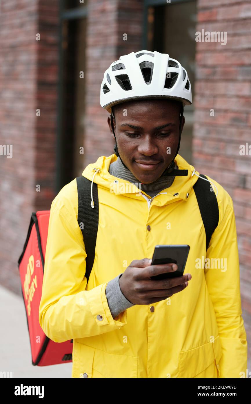 Jeune homme noir dans un casque et une veste jaune regardant l'écran de son smartphone tout en livrant de la nourriture aux clients de la société de service en ligne Banque D'Images
