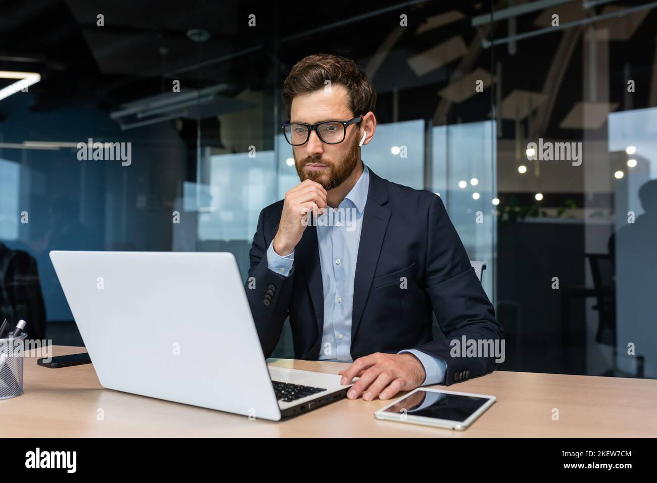 Homme d'affaires senior en lunettes et barbe pense à une décision, l'homme travaille à l'intérieur du bureau et utilise un ordinateur portable au travail, l'investisseur mature dans une combinaison d'affaires est assis à la table Banque D'Images