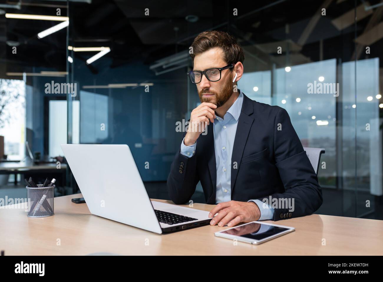 Homme d'affaires senior en lunettes et barbe pense à une décision, l'homme travaille à l'intérieur du bureau et utilise un ordinateur portable au travail, l'investisseur mature dans une combinaison d'affaires est assis à la table Banque D'Images