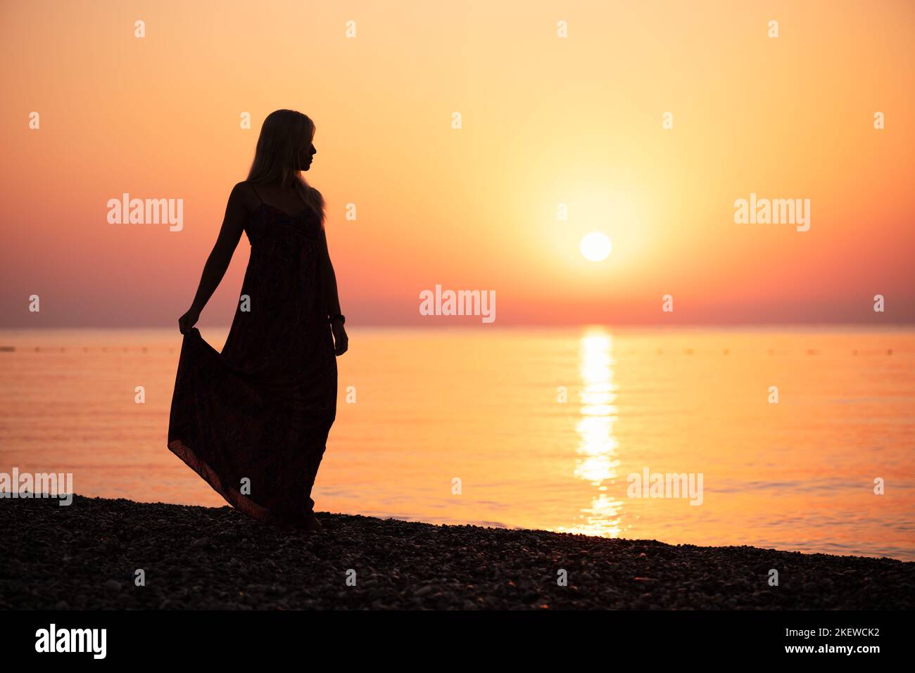 Une femme dans une sundress contemple un coucher de soleil vif au bord de la mer Banque D'Images