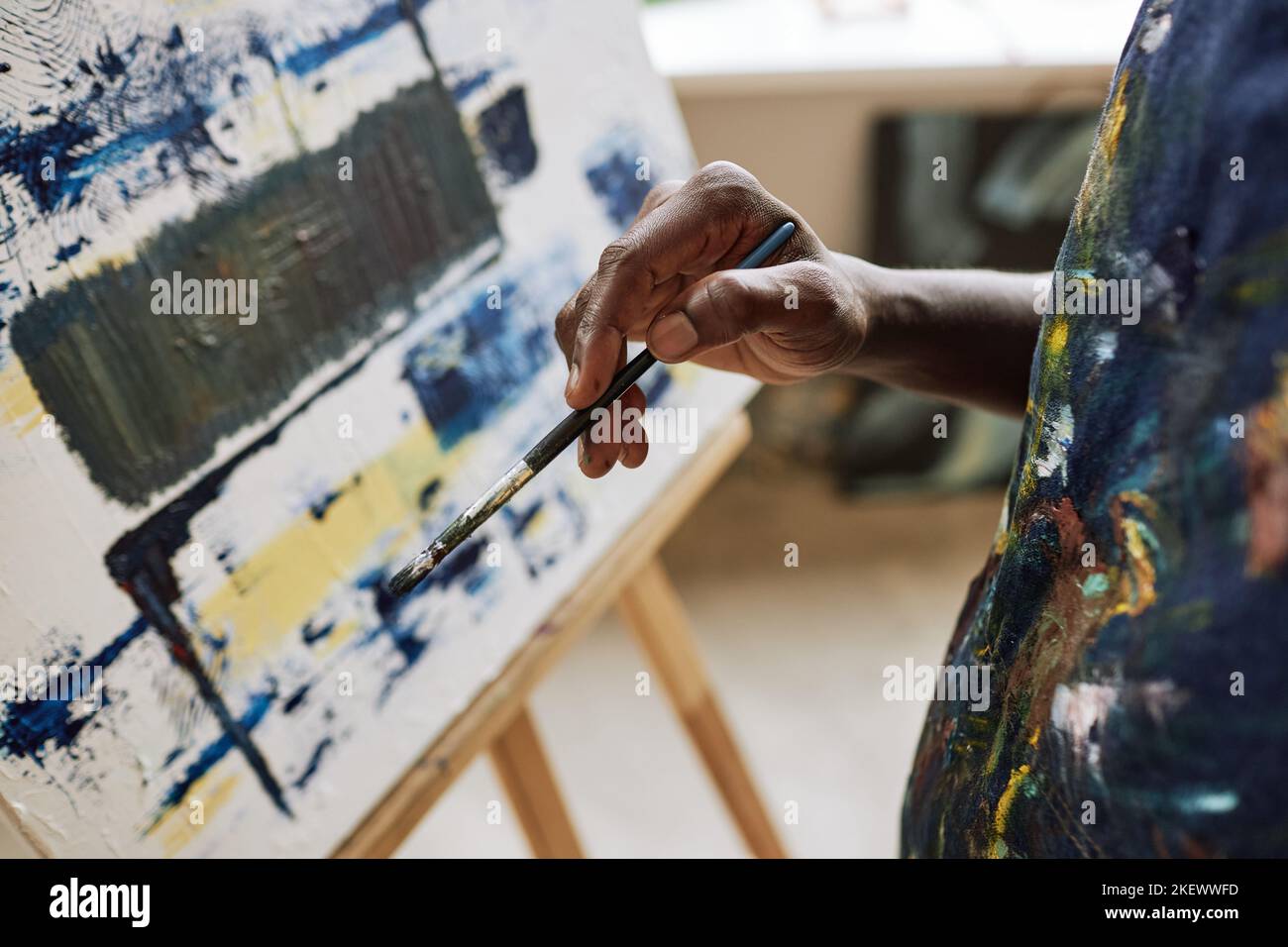 Gros plan de la main d'un jeune peintre afro-américain créatif tenant un pinceau debout devant une toile avec des œuvres d'art inachevées Banque D'Images