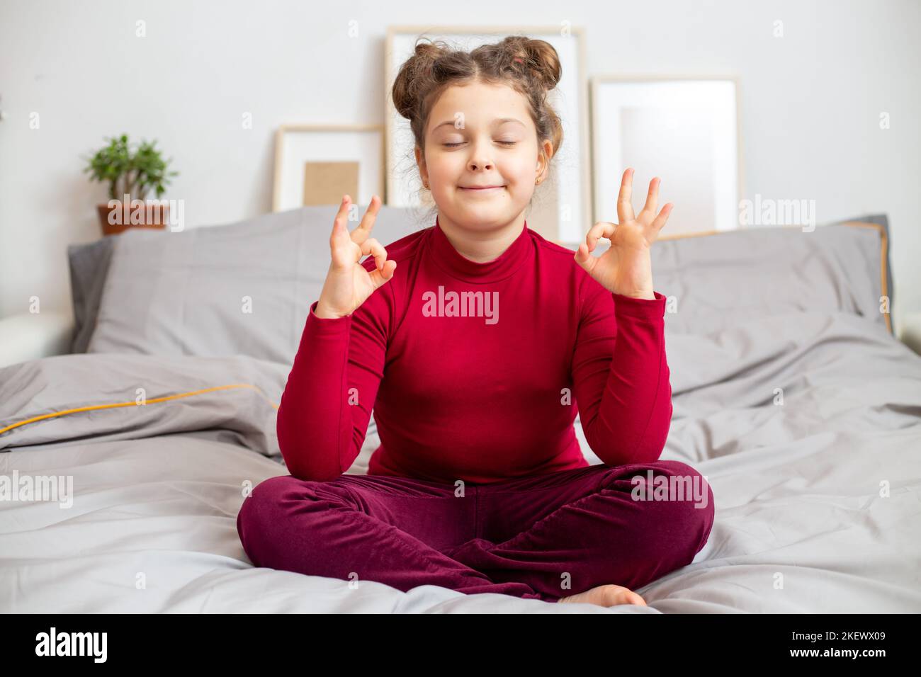 Happy Calm petite fille caucasienne 10s dans un t-shirt bordeaux médite sur un lit gris à la maison. Les enfants se détendent avec le yoga à la méditation pour une vie saine Banque D'Images