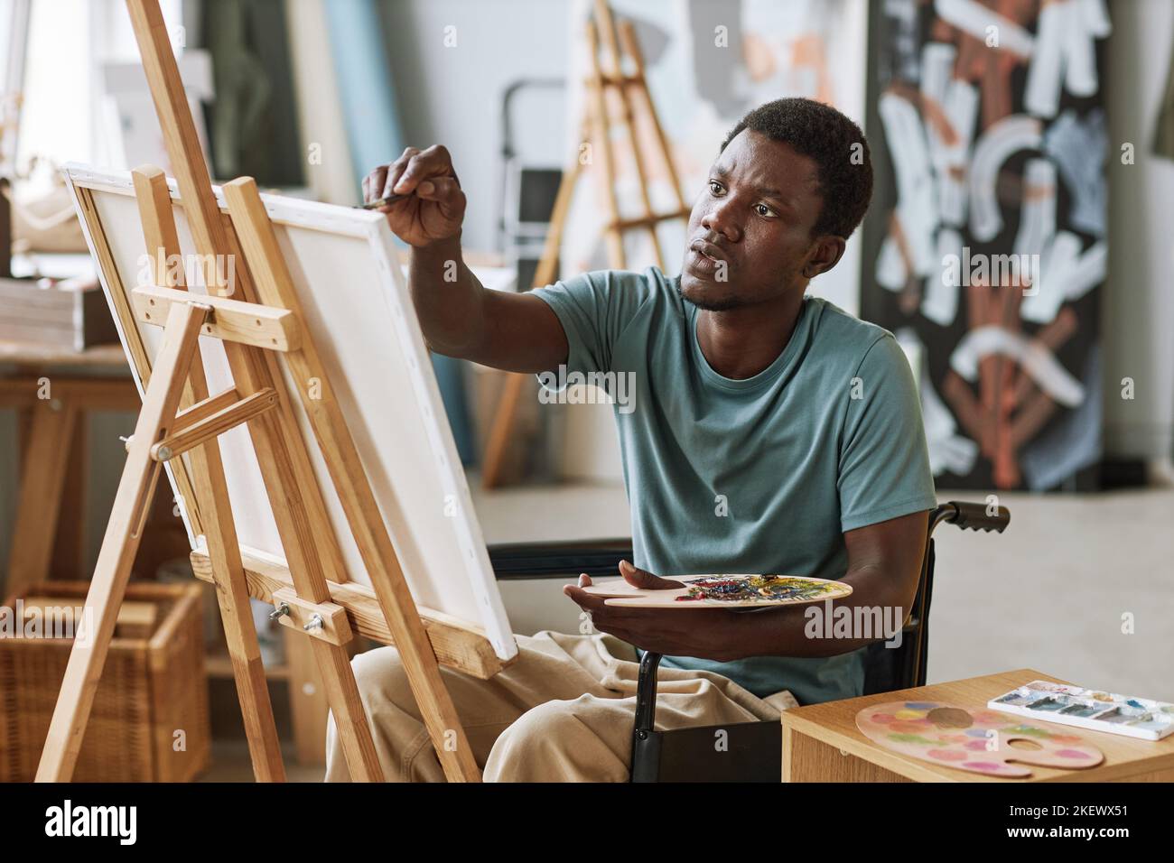 Jeune homme noir en fauteuil roulant avec palette de couleurs dans la main  peinture des œuvres d'art sur toile tout en étant assis devant le chevalet  en studio Photo Stock - Alamy
