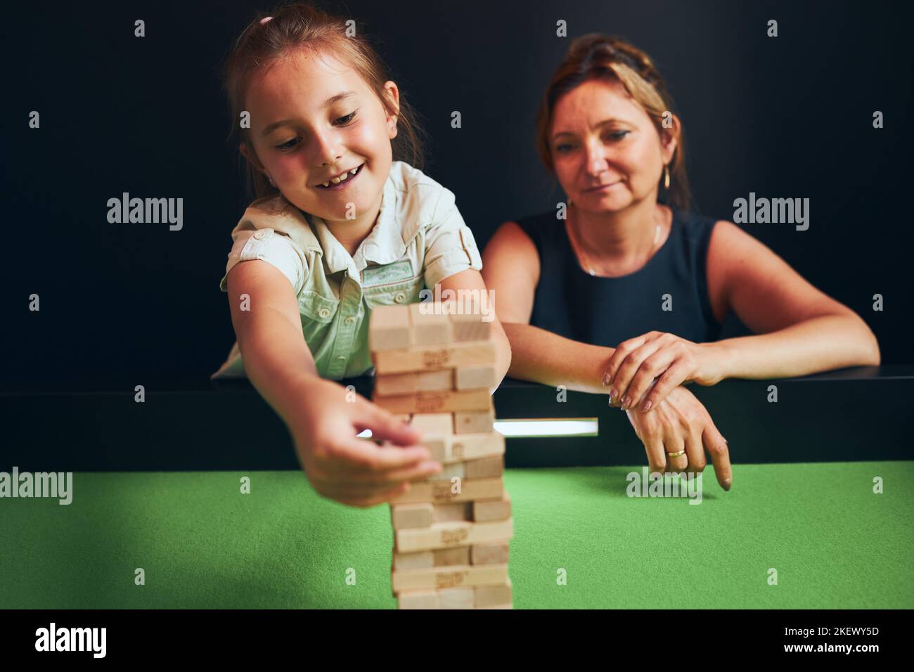 Mère et sa fille jouant à jenga ensemble dans la salle de jeu. Fille en retirant un bloc de la pile et en la plaçant sur le dessus de la tour. Bois empilés bl Banque D'Images