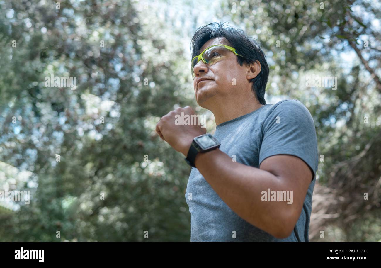 Photo moyenne d'homme latin courant parmi les arbres dans une forêt avec des lunettes et une montre intelligente Banque D'Images