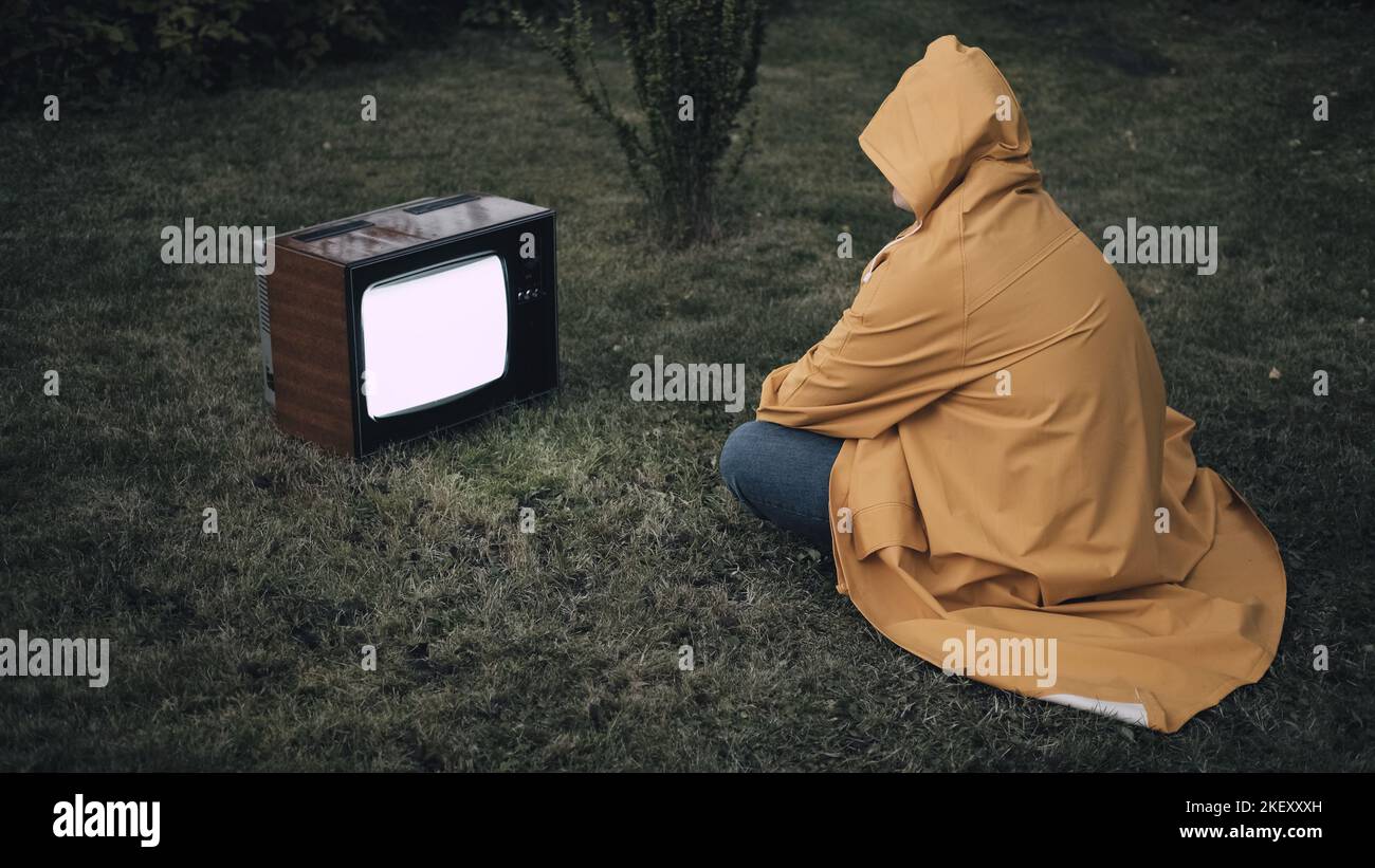 Homme dans un imperméable jaune est assis sur l'herbe et regarde la vieille télé rétro Banque D'Images