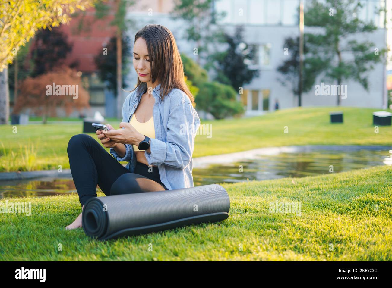 Jolie femme caucasienne assise sur une herbe verte avec un smartphone près du tapis de fitness. Concept de mode de vie sain et exercice sur un air frais. Femme de fitness Banque D'Images