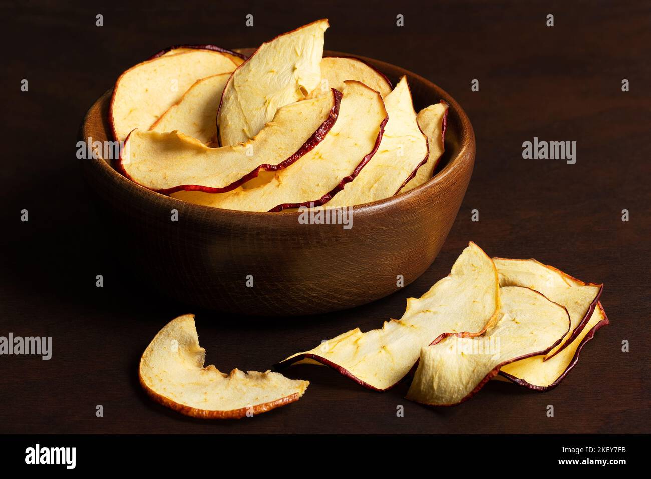 Tranches de pommes séchées dans un bol en bois brun à côté de tranches de pommes séchées déversées sur fond de bois foncé. Banque D'Images