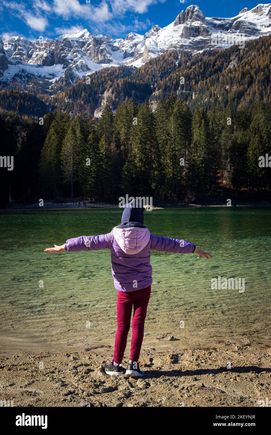 Enfant fille debout à bras ouverts près des eaux cristallines du lac Tovel, Trentin-Haut-Adige, Italie. Vue sur l'automne avec des montagnes enneigées dans la ba Banque D'Images