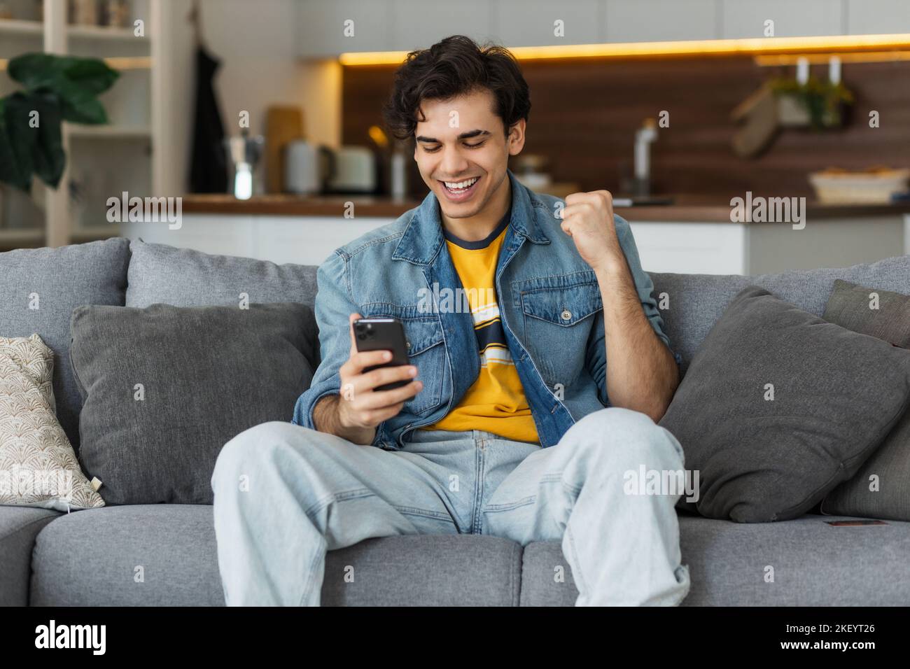 jeune homme caucasien excité célébrant le succès. Un jeune homme lisant de bonnes nouvelles en utilisant un téléphone portable assis au canapé Banque D'Images