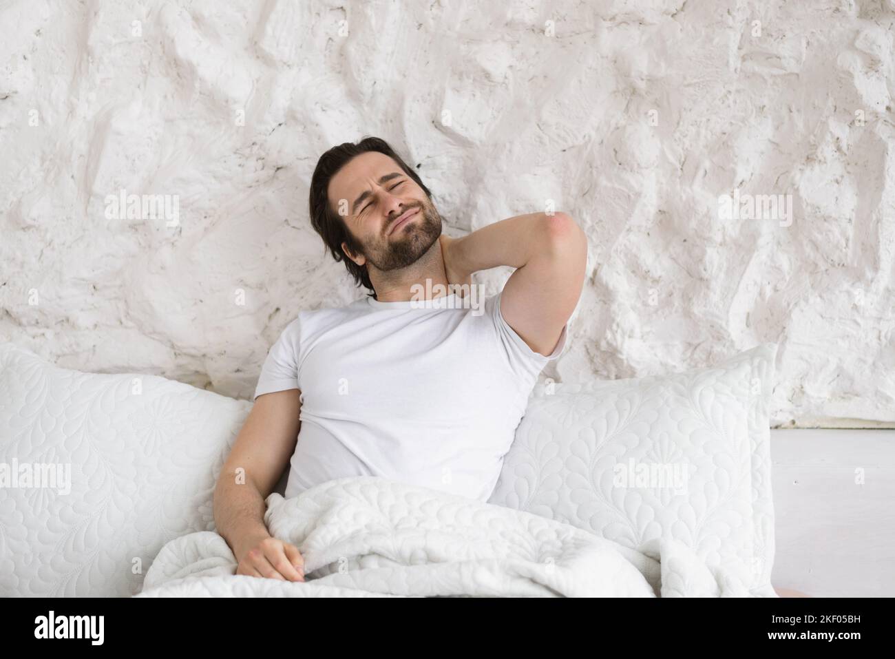 Triste homme de race blanche millénaire désespéré avec un chaume maque son cou sur un lit blanc aux couvertures et souffre de mal Banque D'Images