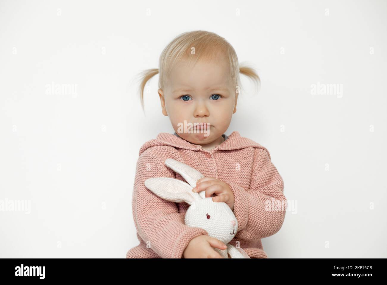 Triste bébé fille jouer avec animal bourré isolé sur blanc. Un tout-petit avec un visage troublé tient dans les mains le lapin en peluche. Enfant blond aux cheveux en rose Banque D'Images