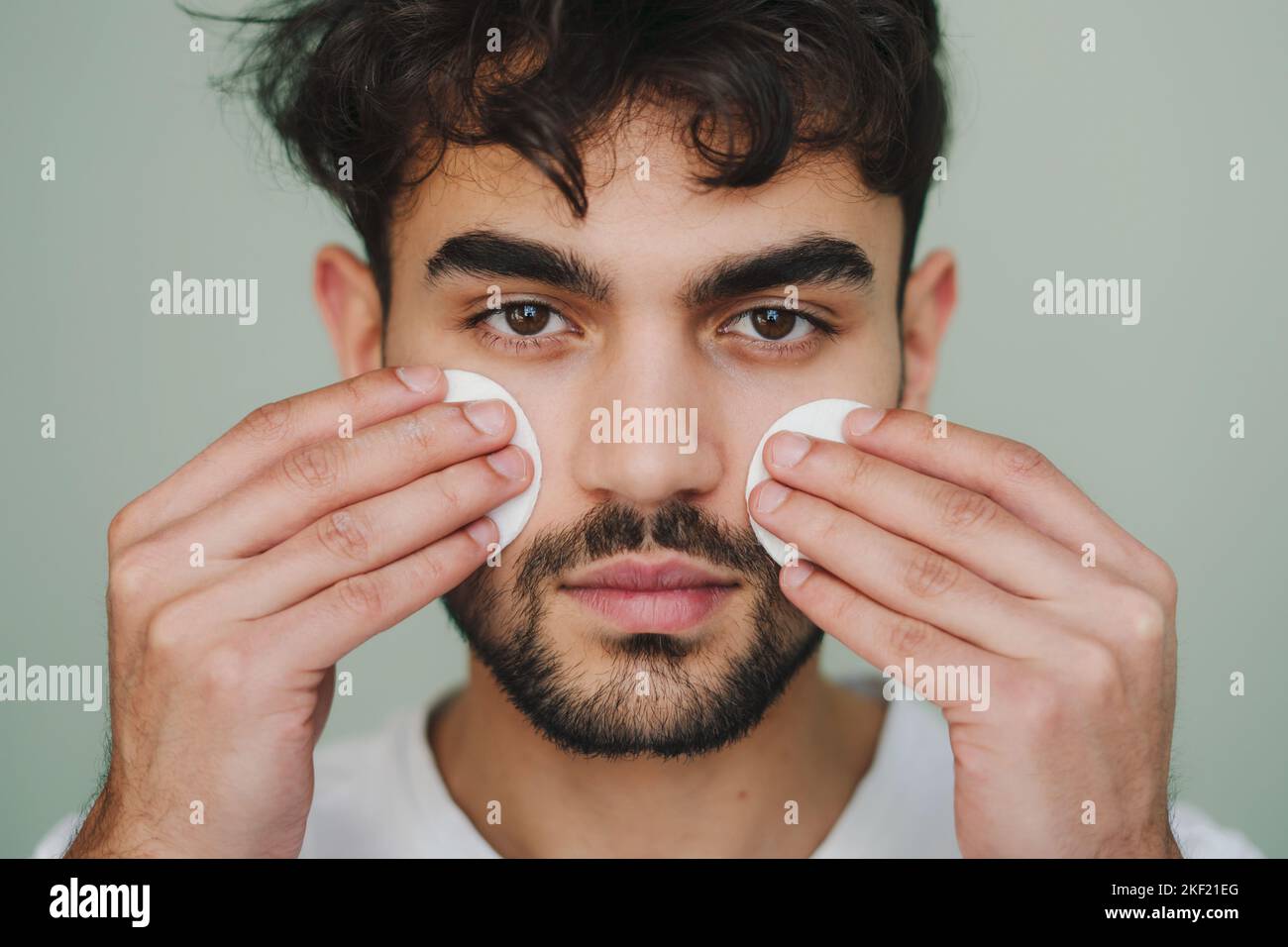 Portrait de beauté d'un jeune homme confiant faisant le nettoyage du visage de routine du matin avec des tampons de coton, isolé sur fond vert. Soins de santé. Beauté Banque D'Images