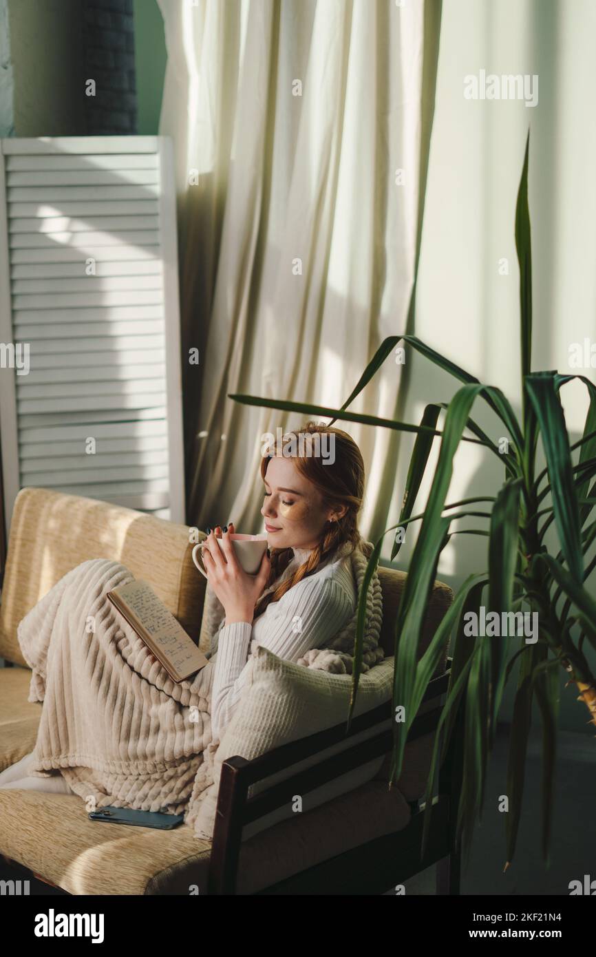 Jeune femme gaie avec des taches sous les yeux tenant une tasse de café, assis avec un livre sur un canapé. Soins du corps. Soins de beauté. Soins des yeux. Banque D'Images