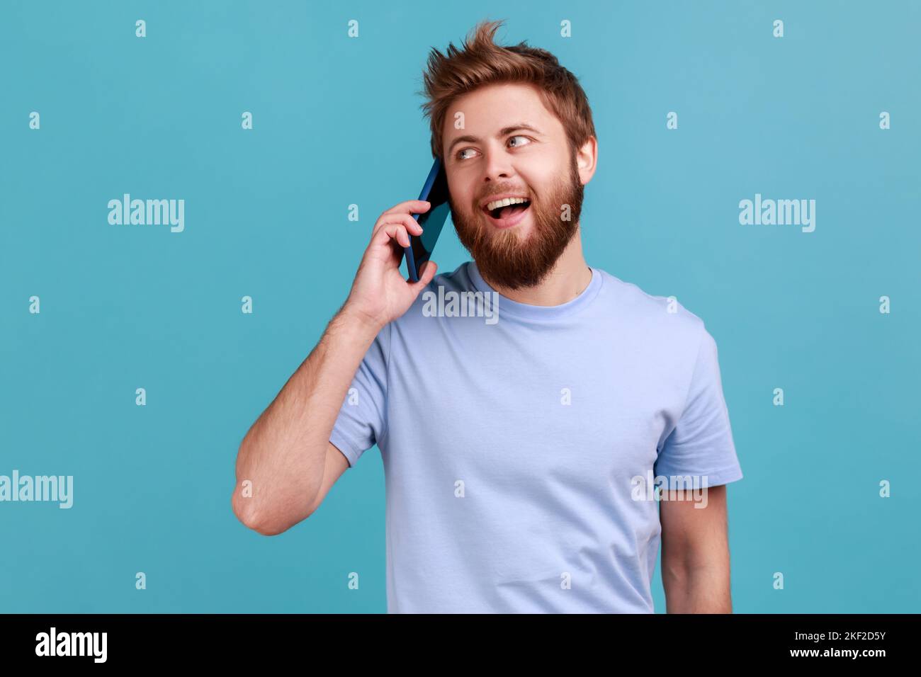Portrait d'un bel homme barbu excité debout, parlant smartphone, passer un appel et avoir une conversation agréable avec un ami ou une femme. Studio d'intérieur isolé sur fond bleu. Banque D'Images